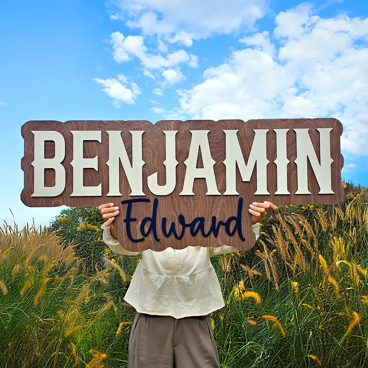 Person holding a wooden sign with 'Benjamin Edward' against a blue sky and grassy field background