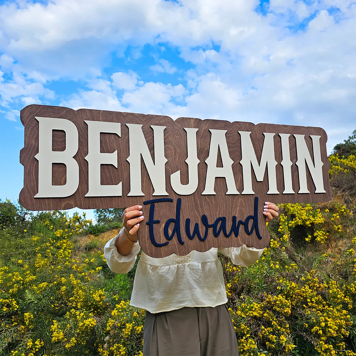 Person holding a wooden sign with 'Benjamin Edward' against a blue sky and yellow flowers.